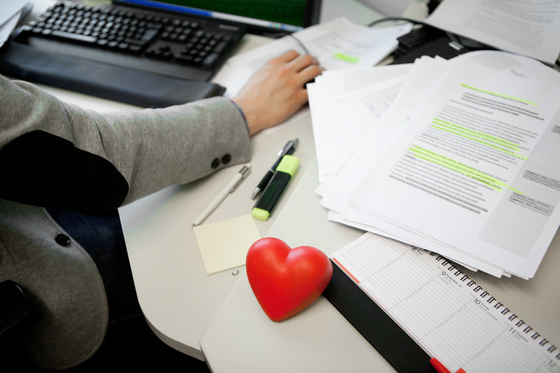 työpöydällä läppäri, papereita ja punainen sydän, kuvassa myös käsi